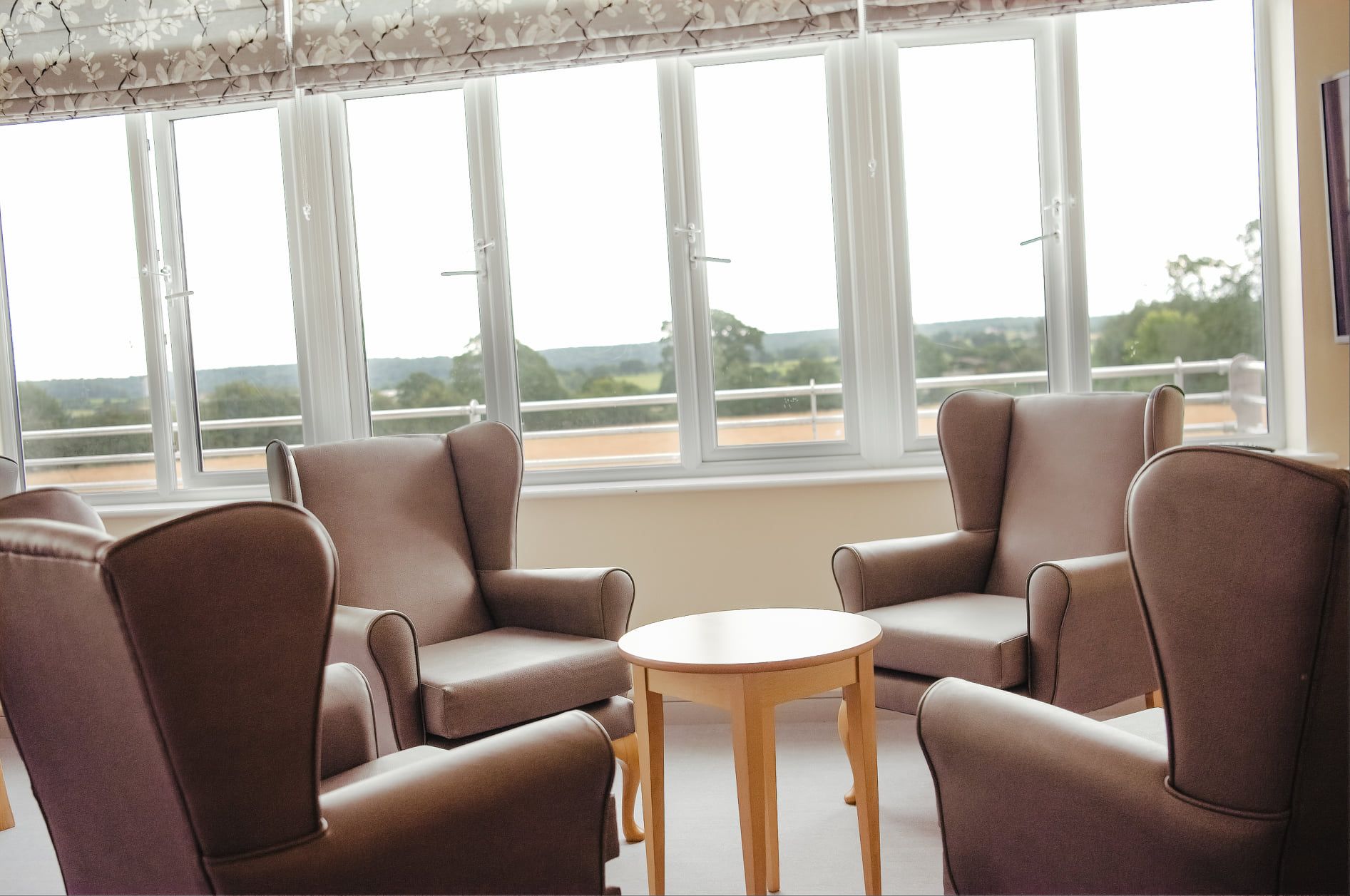 Wing-back armchairs around a small table, facing a wide window over the fields.